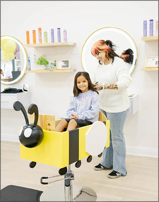 Child sitting in a bee-themed chair getting a haircut in a salon.