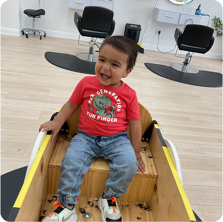 Child in a red shirt sitting inside a wooden toy boat in a salon setting