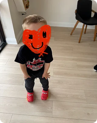 Child wearing a black t-shirt with graphics, gray pants, and red shoes, standing on a wooden floor.