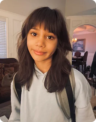 Young girl with a backpack indoors, smiling at the camera.