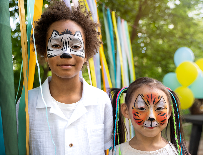 Two children with animal face paint standing in front of colorful ribbons and balloons.